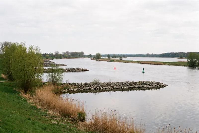 Breiter, ruhiger Fluss mit einer Reihe Steinbuhnen am Ufer und zwei Fahrwassertonnen in der Mitte, dahinter weitet sich das Wasser unter grauem Himmel