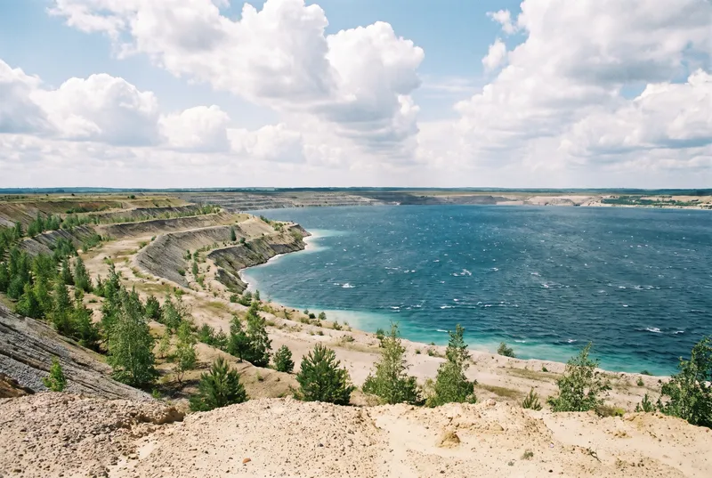 Junger Tagebausee von oben gesehen, die Böschung fällt in Sandterrassen mit jungen Kiefern ab, davor weites blaues Wasser mit Wellenkämmen unter Quellwolken