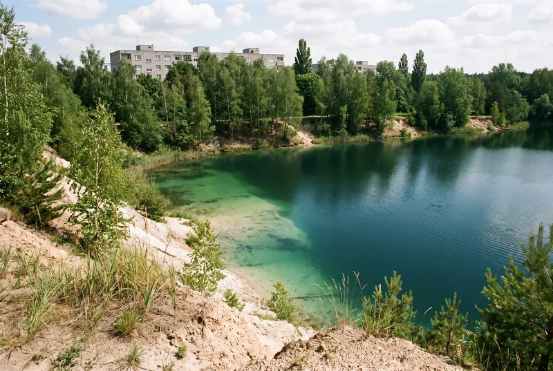 Ehemalige Kiesgrube im Frühsommer von der sandigen Uferkante aus, unter dem klaren Wasser fällt die helle Kiesflanke steil ab, ringsum Birken und Kiefern, über den Baumwipfeln die Dächer eines Plattenbaus.