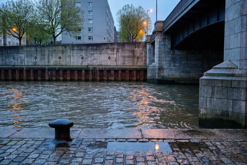 Stadtkanal in der blauen Stunde, vorn nasses Kopfsteinpflaster mit einem eisernen Poller, gegenüber eine Betonwand über rostiger Spundwand, rechts die Granitpfeiler einer Straßenbrücke, Laternenlicht liegt auf dem Wasser.