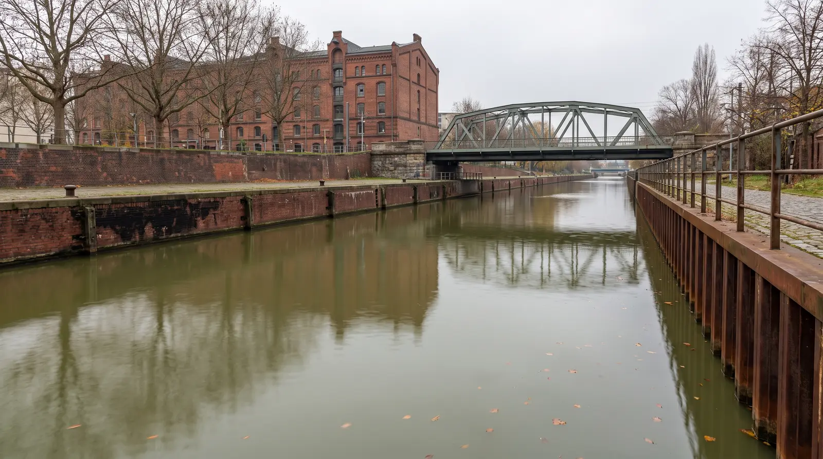 Stadtkanal an einem grauen Herbsttag, links eine Backsteinmauer mit Pollern vor einem alten roten Speichergebäude, rechts eine rostige Spundwand. Über dem ruhigen olivgrünen Wasser spannt sich eine grüne Stahlfachwerkbrücke.