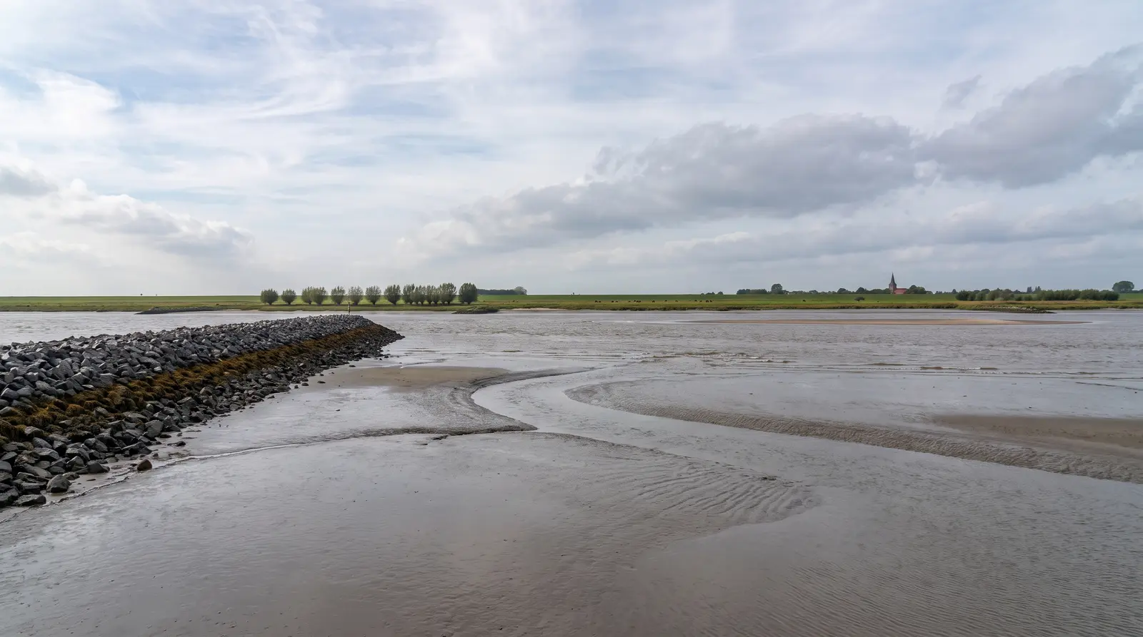 Tideweser bei Niedrigwasser, eine Steinschüttung zieht von links in die freigefallenen Schlickflächen, davor ein flacher Priel mit Rippelmarken im nassen Sand. Dahinter die flache Marsch mit Deich, einer Reihe Kopfweiden und einem Kirchturm unter hohem, bewölktem Himmel