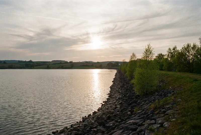 Abendsonne über einem gebauten See, eine lange Steinschüttung führt am Damm entlang ins Bild, dahinter frisch belaubte Birken und flache Felder