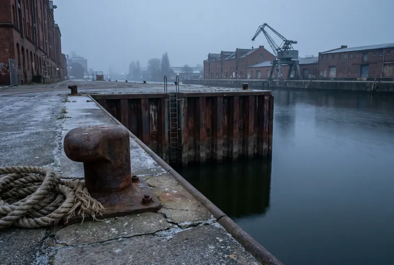 Hafenbecken im grauen Winterlicht, vom Kai aus fällt eine rostige Spundwand senkrecht ins dunkle, stille Wasser, davor ein Poller mit aufgeschossenem Tau, dahinter Backsteinschuppen und ein alter Hafenkran