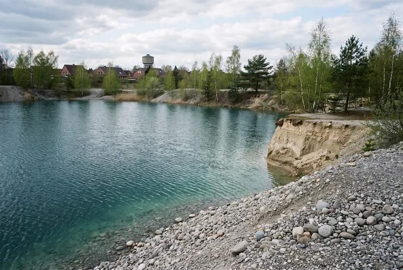 Ausgekieste Grube am Stadtrand im Frühjahr, eine helle Kiesbank fällt steil ins klare türkise Wasser, rechts eine abgebrochene Sandkante, am Ufer Birken und Kiefern, dahinter Hausdächer und ein Wasserturm