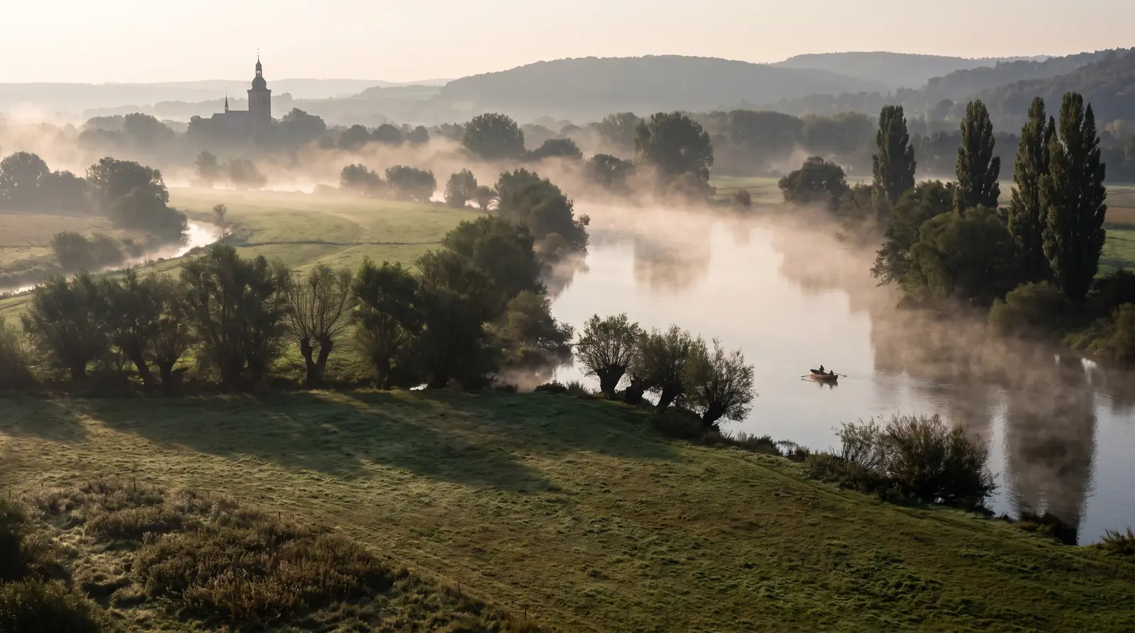 Flusstal im Morgennebel, ein Boot mit Angler auf dem Wasser, Weiden und Wiesen am Ufer, Kirchturm am Horizont