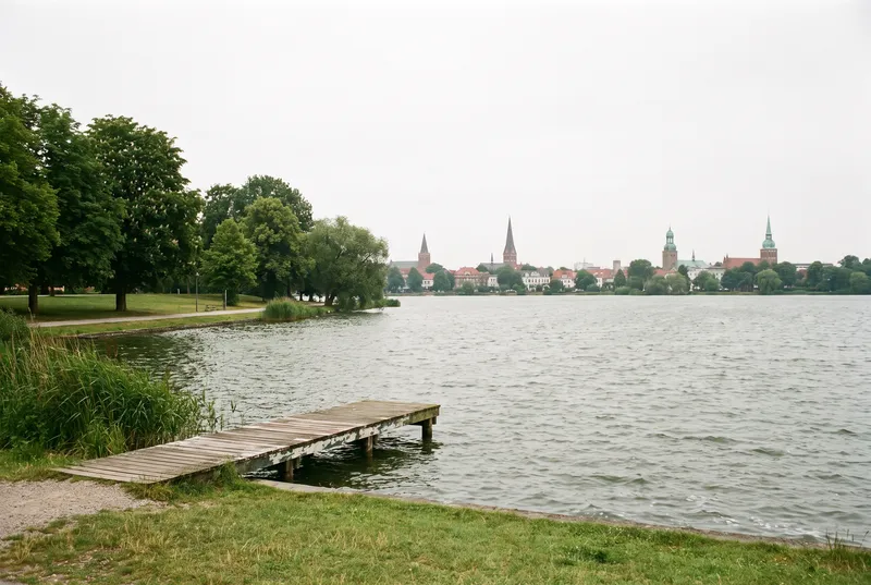 Weite Wasserfläche eines Stadtsees an einem bedeckten Sommermittag, im Vordergrund ein Holzsteg an der Schilfkante und eine Liegewiese, am gegenüberliegenden Ufer die Türme der Innenstadt