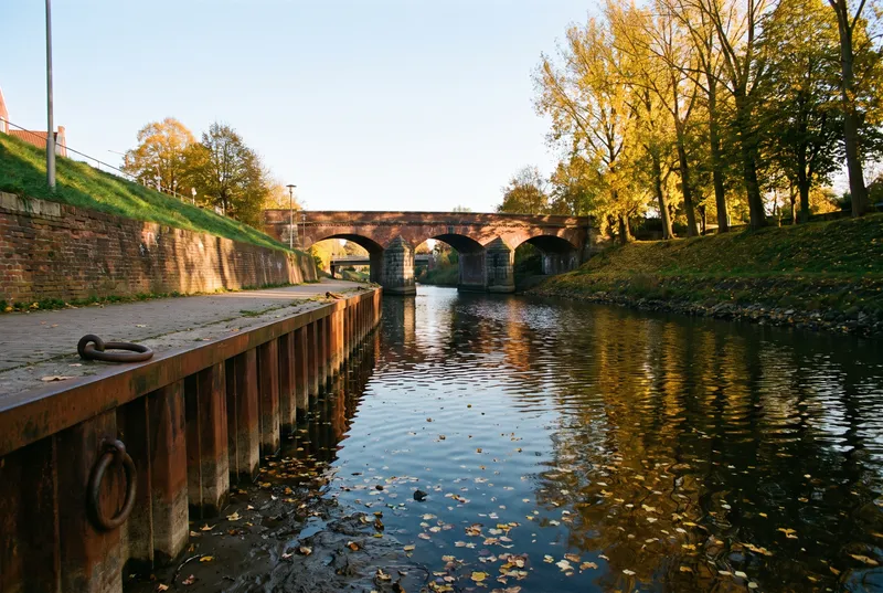 Gestauter Flusslauf an einem Herbstnachmittag, links Spundwand mit Ringen und eine Backsteinmauer unter dem Uferweg, geradeaus eine Ziegelbogenbrücke, auf dem dunklen Wasser treibt Laub