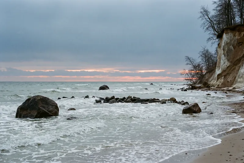Brandung der offenen Ostsee im ersten Morgenlicht, große Findlinge und ein Steinriff im Wasser, rechts ein helles Kliff aus Kreide und Geschiebemergel mit kahlen Buchen darüber