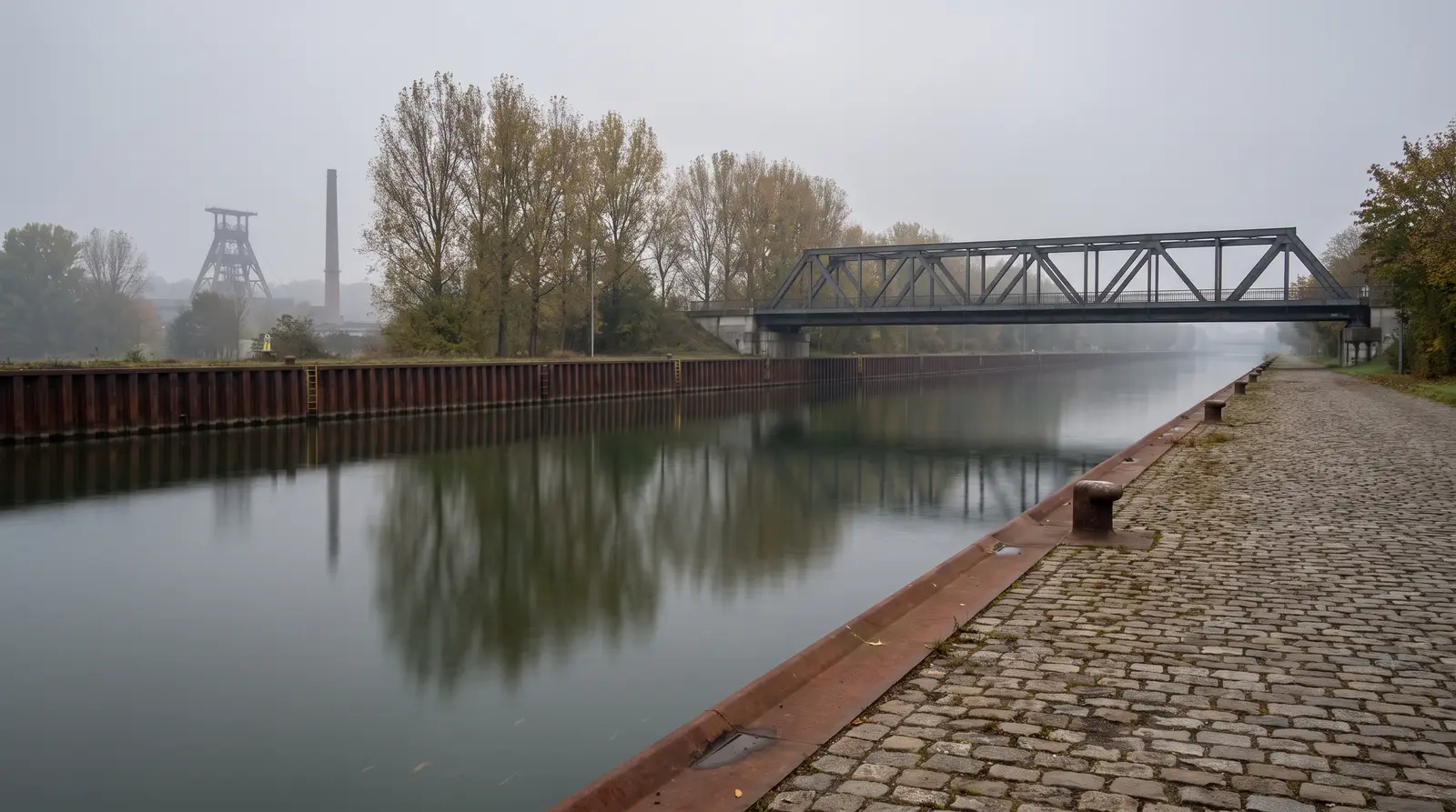 Schnurgerader Schifffahrtskanal im Herbstdunst, am linken Ufer eine rostige Stahlspundwand, rechts ein grob gepflasterter Treidelweg mit Pollern, in der Ferne eine Stahlfachwerkbrücke und die Silhouette eines Förderturms mit Industrieschornstein