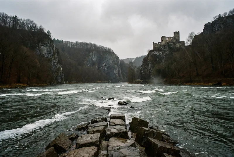 Steinbuhne aus nassen Blöcken ragt in den aufgewühlten Strom, an beiden Seiten steile Felsen und kahler Wald, oben rechts eine Burgruine auf dem Fels
