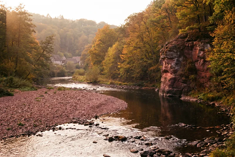 Flussbiegung im Herbstlicht, links eine breite Kiesbank aus rötlichen Steinen mit flacher Rausche, rechts ein tiefer Kolk unter einer roten Felswand
