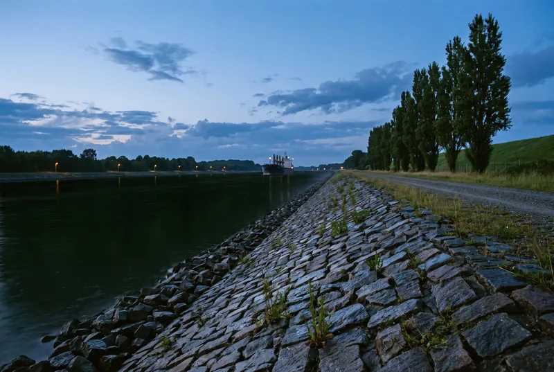 Eine steile Böschung aus groben Granitsteinen zieht sich als Fluchtlinie am schnurgeraden Kanal entlang, oben ein Betriebsweg mit Pappelreihe, in der Ferne die Silhouette eines Frachters in der blauen Stunde