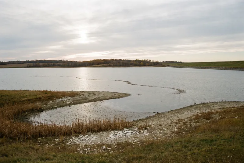 Weite ruhige Stauseefläche unter tief stehender Sonne hinter dünnen Wolken, vorne eine flache Bucht mit Kies, trockenem Schilf und abgesunkener Wasserlinie, rechts ein begrünter Damm