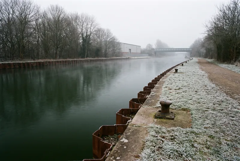 Kanalisierter Fluss im Winter, rechts eine rostige Spundwand und eine Betonkante mit eisernen Pollern, dahinter ein raureifweißer Grasstreifen mit Kiesweg, im Dunst flussab eine Werkshalle und eine Stahlbrücke