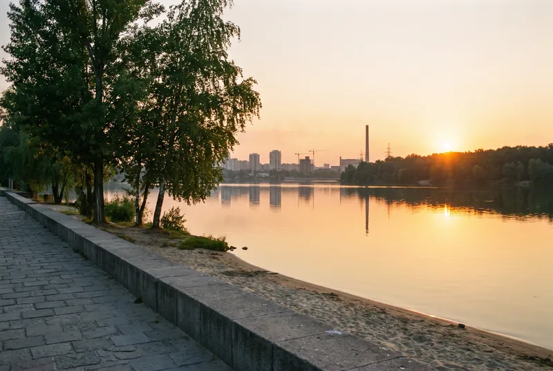 Gepflasterte Uferpromenade mit Steinkante und schmalem Sandstreifen an einem großen See, gegenüber die Silhouette einer Stadt im Abendlicht