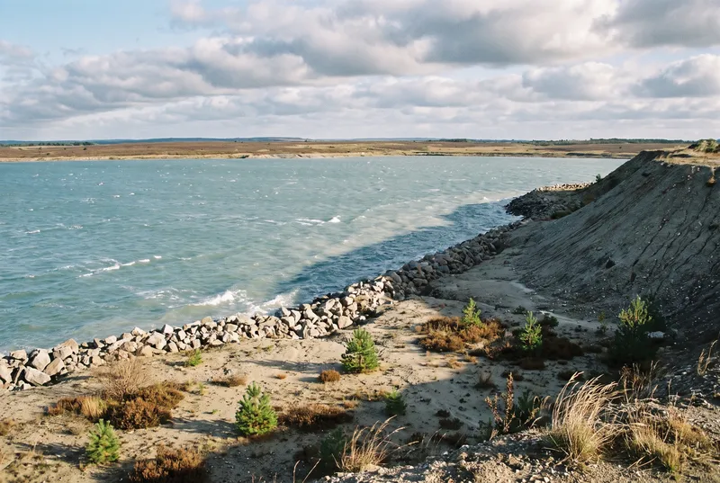 Junger Tagebausee mit weißen Schaumkronen, unten am Ufer eine grobe Steinschüttung vor roher Sandböschung mit kleinen Kiefern