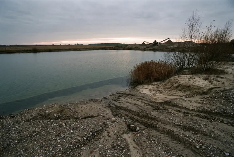 Junger Kiessee in flachem Ackerland kurz nach Sonnenuntergang, Sandufer mit Reifenspuren im Vordergrund, am anderen Ufer Kiesberge und ein Förderband
