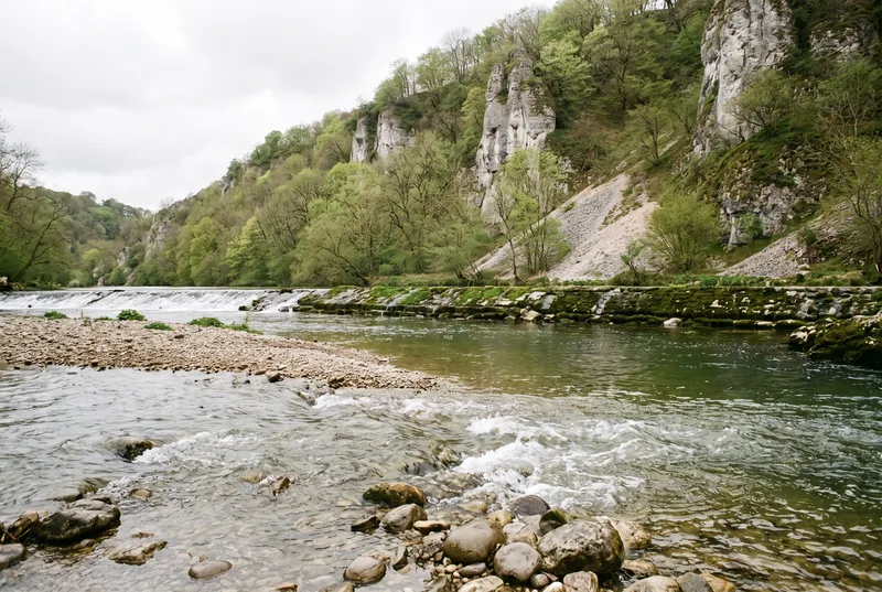 Frei fließender Fluss über einer Kiesbank, im Vordergrund ein Rauschen mit weißem Wasser, dahinter ein flaches Wehr und helle Kalkfelsen über dem Ufer