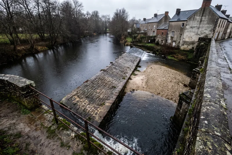 Altes Steinwehr in einem Fluss, von einer Brücke aus gesehen, darunter eine helle Kiesbank, am rechten Ufer die Rückseiten alter Häuser im Regen